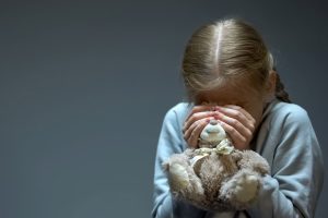 Dziewczynka kuli się ze strachu, zasłania oczy trzymanemu w dłoniach pluszowemu misiowi, za którym ukrywa swoją twarz. Ilustracja do artykułu o problemach wychowawczych związanych z zaburzeniami lękowymi